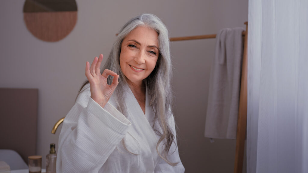 Happy senior mature lady Caucasian old woman female in bathrobe in bathroom showing okay perfect shower gel cosmetic anti-wrinkle procedure show ok gesture satisfied with anti-aging body skin care